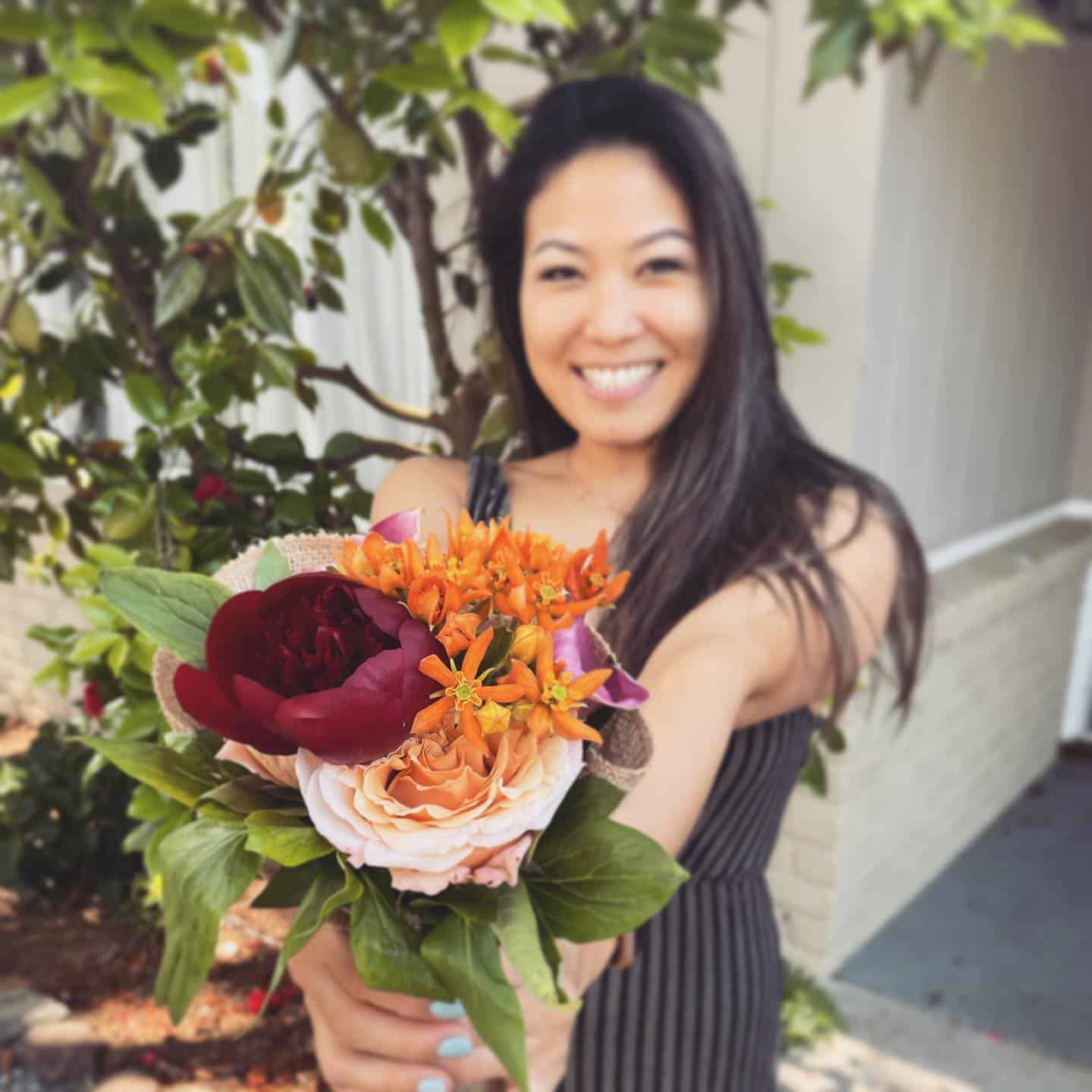 A woman standing outdoors holds a bouquet of colorful flowers toward the camera, smiling. Green foliage and a building are visible in the background.