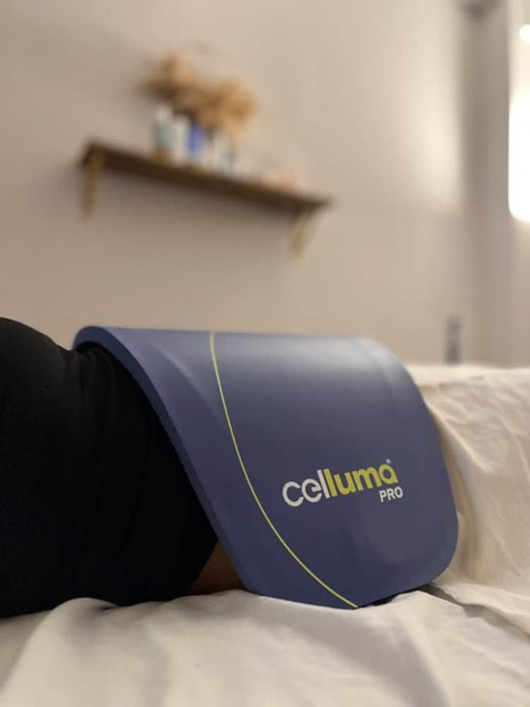 A Celluma PRO light therapy panel is placed on a person's body in a softly lit treatment room with shelves on the wall.