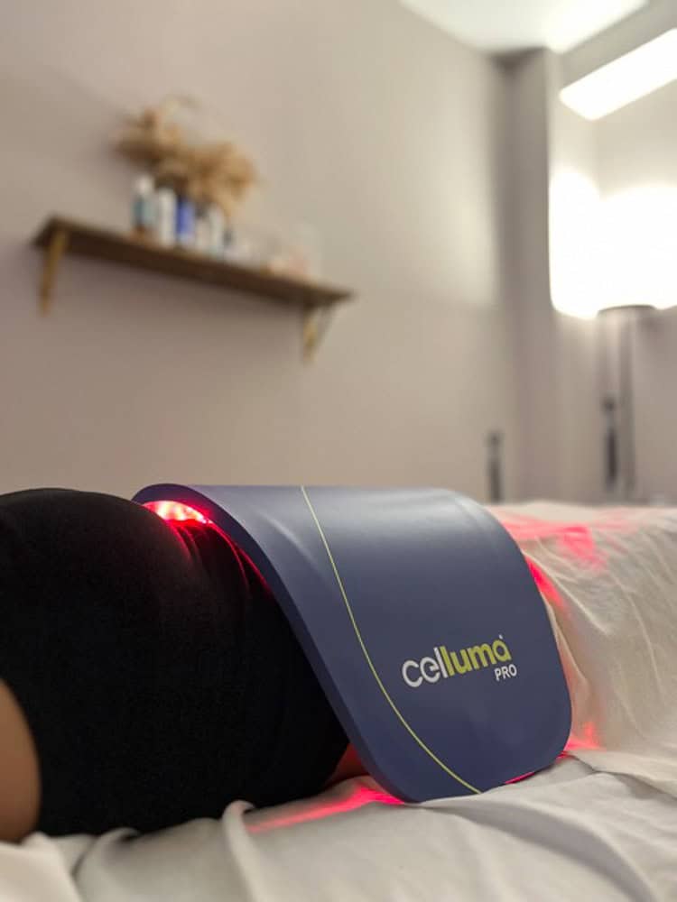 A Celluma PRO light therapy panel is placed on a person's body in a softly lit treatment room with shelves and framed certificates on the wall.