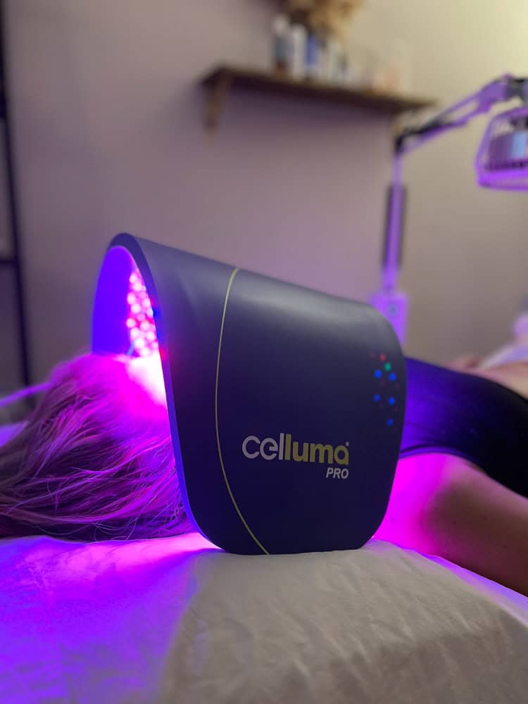 A Celluma PRO light therapy panel is placed on a person's body in a softly lit treatment room with shelves and framed certificates on the wall.